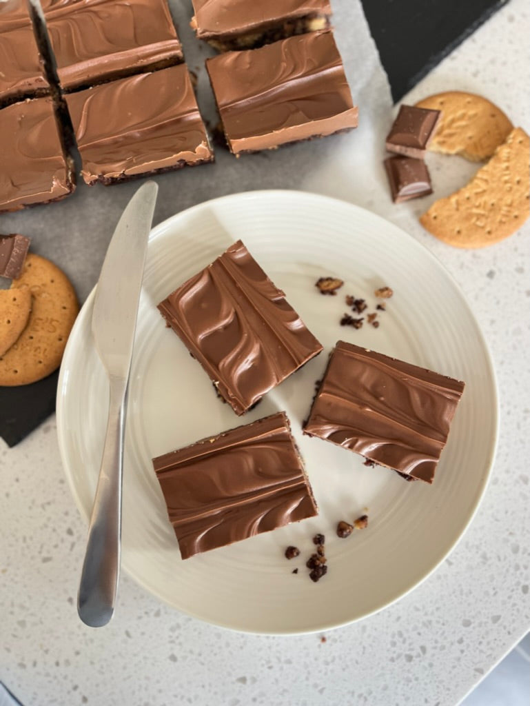 Delicious Tiffin pieces topped with milk chocolate served on a white plate, surrounded by digestive biscuits.